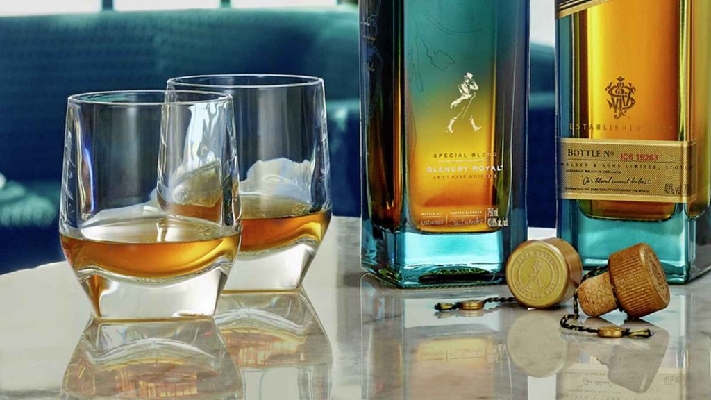 Imagem mostra dois copos baixos com whisky, ao lado de duas garrafas de Johnnie Walker Blue Label e Ghost & Rare Glenury Royal.