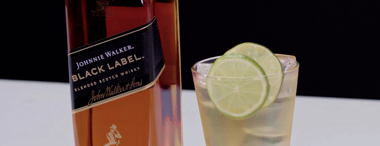 Drink Black Tonic Highball, em copo alto com gelo e fatia de limão, ao lado de garrafa de Johnnie Walker Black Label.