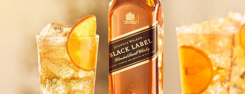 Imagem mostra copos de drink highball com bastante gelo e fatias de laranja. Ao centro, uma garrafa de whisky Johnnnie Walker Black Label.