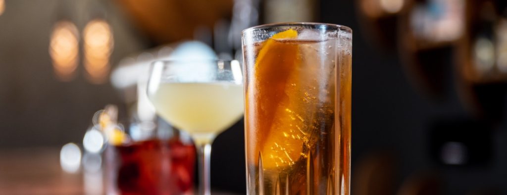 Imagem mostra copo highball com drink de cor dourada, uma pedra de gelo e um pedaço de casca de laranja. Ao fundo, uma taça martini com drink de cor amarelo claro e um negroni servido em copo baixo.